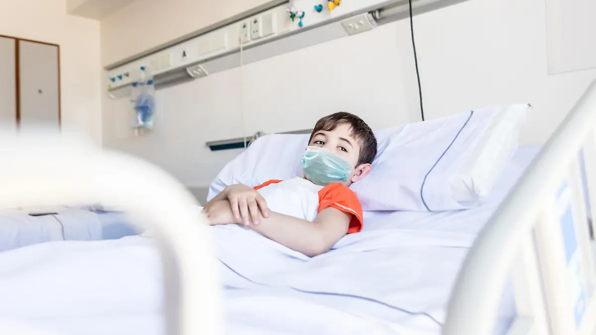 Pensive little boy lying down on hospital bed looking away with sadness wearing a protective facemask - Pandemic lifestyles