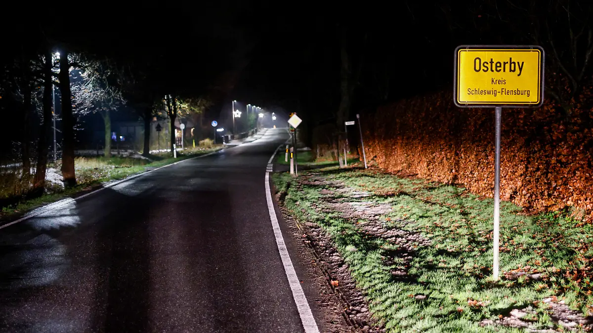 PRODUKTION - 19.12.2022, Schleswig-Holstein, Osterby: Das Ortschild von Osterby und die Strassenbeleuchtung entlang der Hauptstraße. Das Dorf spart Strom mit einer außergewöhnlichen "Licht an"-Idee. In Osterby wird die Straßenbeleuchtung samt Weihnachtslichtern von 22:30 Uhr bis 05:00 Uhr komplett ausgestellt. Diese Maßnahme soll helfen, Energiekosten einzusparen. Damit aber die Anwohner oder auch Besucher während dieser Zeit nicht im dunkeln gehen oder den Hund ausführen müssen, gibt es vor Ort einen ganz besonderen SMS-Service. Das Wort "Licht" an eine dafür vorgesehene Nummer schreiben und in wenigen Sekunden gehen alle Laternen für rund 15 Minuten an. Foto: Axel Heimken/dpa +++ dpa-Bildfunk +++