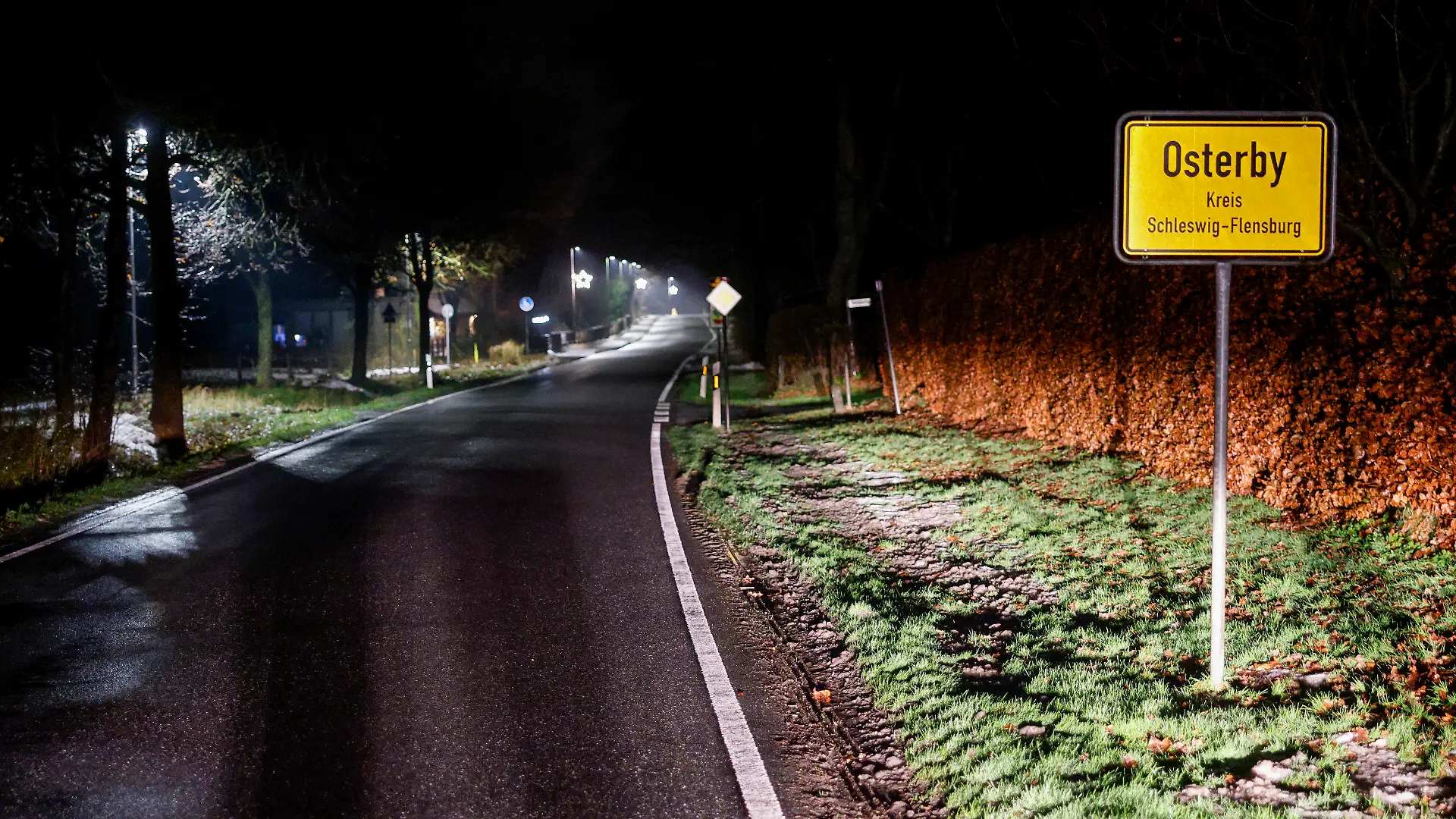 PRODUKTION - 19.12.2022, Schleswig-Holstein, Osterby: Das Ortschild von Osterby und die Strassenbeleuchtung entlang der Hauptstraße. Das Dorf spart Strom mit einer außergewöhnlichen "Licht an"-Idee. In Osterby wird die Straßenbeleuchtung samt Weihnachtslichtern von 22:30 Uhr bis 05:00 Uhr komplett ausgestellt. Diese Maßnahme soll helfen, Energiekosten einzusparen. Damit aber die Anwohner oder auch Besucher während dieser Zeit nicht im dunkeln gehen oder den Hund ausführen müssen, gibt es vor Ort einen ganz besonderen SMS-Service. Das Wort "Licht" an eine dafür vorgesehene Nummer schreiben und in wenigen Sekunden gehen alle Laternen für rund 15 Minuten an. Foto: Axel Heimken/dpa +++ dpa-Bildfunk +++
