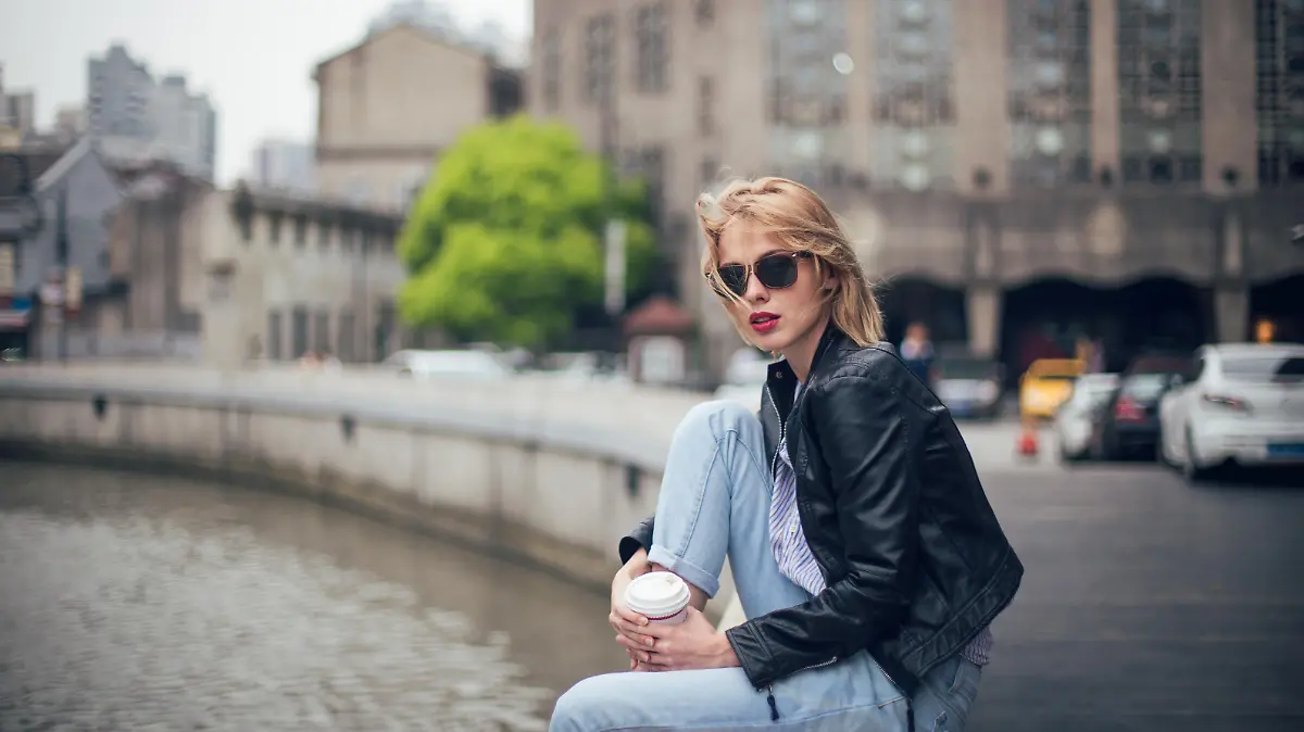 Beautiful young woman in leather jacket sitting by the river and drinking takeaway coffee.