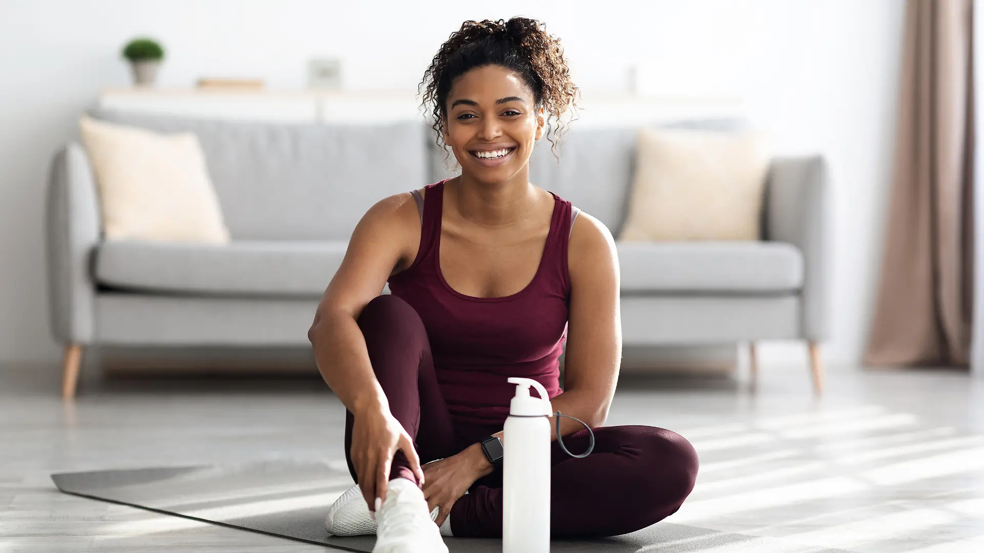 Positive sporty millennial black lady in beautiful sportswear and white sneakers sitting on fitness mat with bottle of water and smiling, having break while exercising at home, copy space