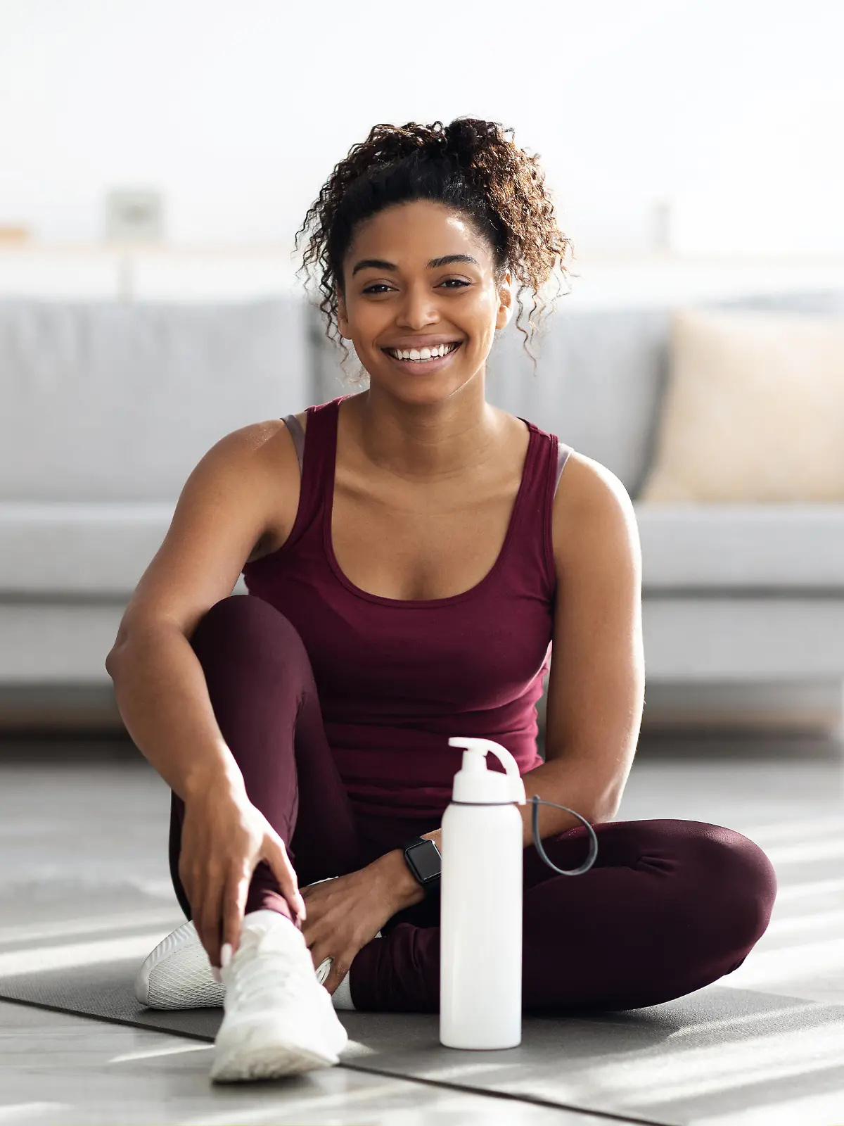 Positive sporty millennial black lady in beautiful sportswear and white sneakers sitting on fitness mat with bottle of water and smiling, having break while exercising at home, copy space