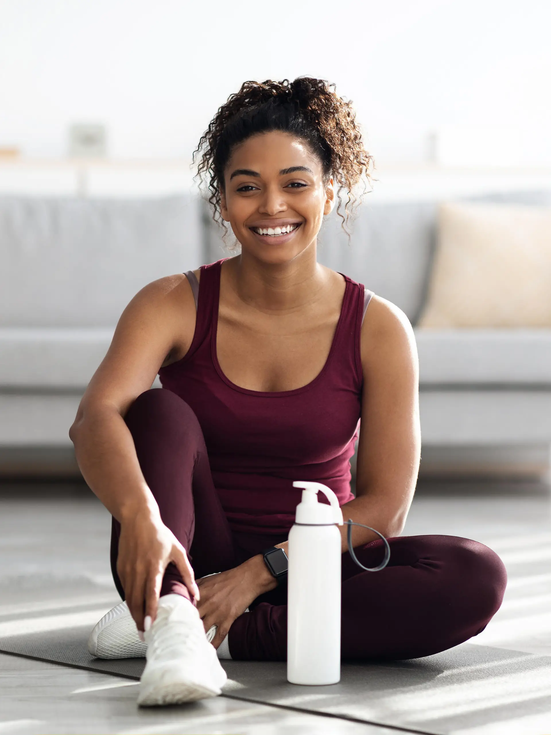 Positive sporty millennial black lady in beautiful sportswear and white sneakers sitting on fitness mat with bottle of water and smiling, having break while exercising at home, copy space