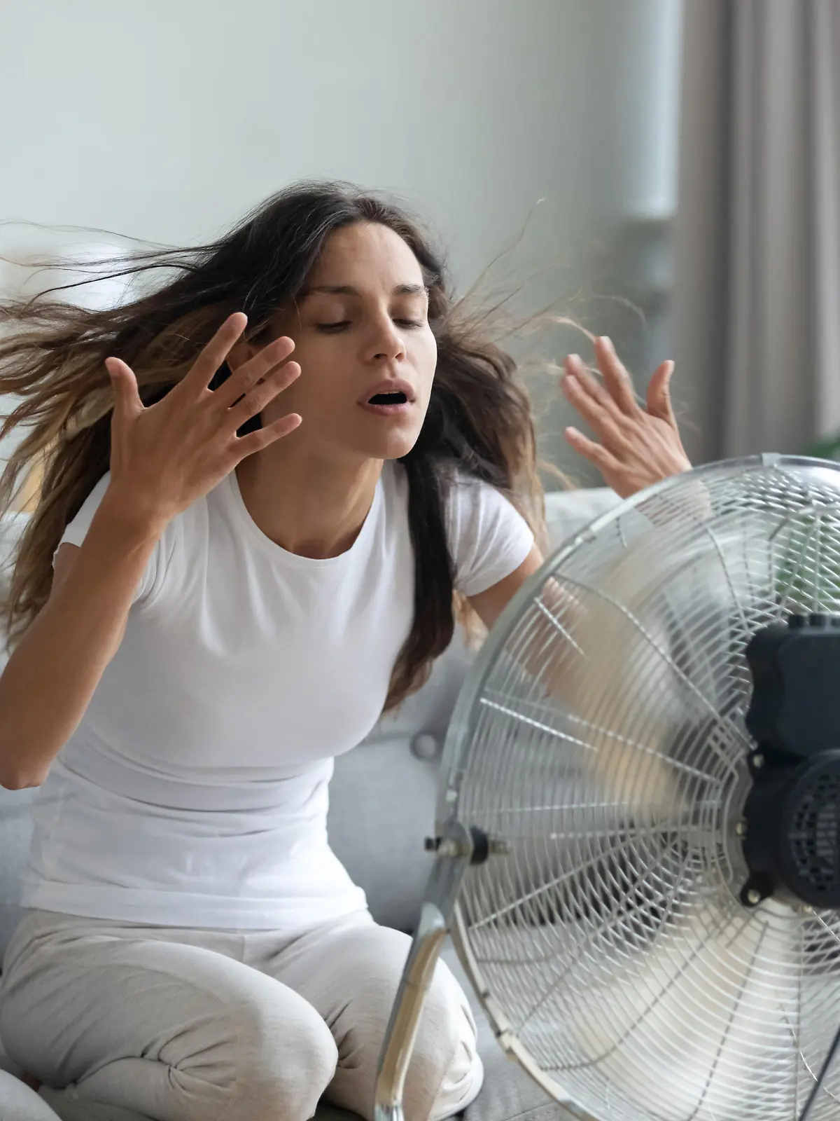 Eine junge Frau sitzt in ihrem Wohnzimmer auf der Couch und hat einen großen Ventilator vor sich stehen.