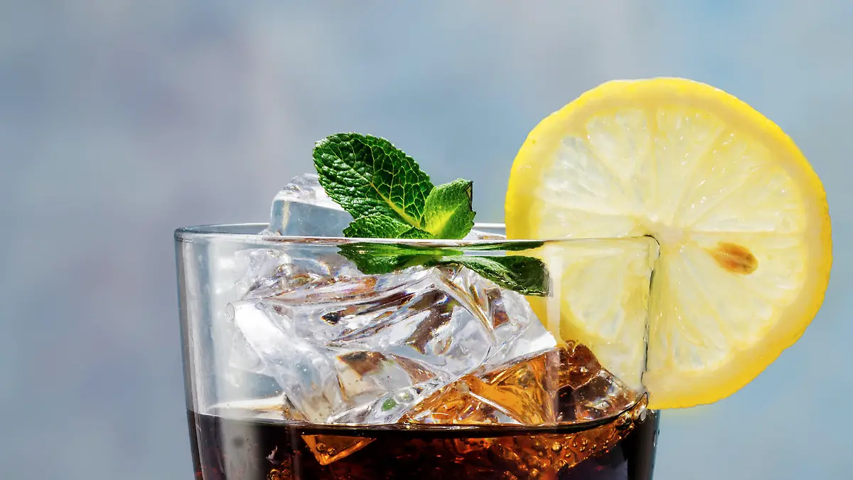 glass of cola or ice tea with ice cubes, lemon slice and peppermint garnish, against a blue wall