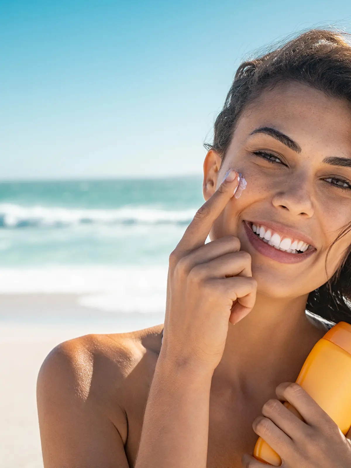 Beautiful young woman at beach applying sunscreen on face and looking at camera. Beauty latin girl enjoying summer holiday while applying suntan lotion at sea. Portrait of happy woman with healthy skin applying sunblock on cheek.