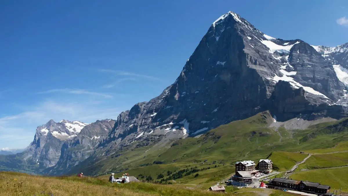 Die Eiger Nordwand an einem klaren sonnigen Tag, mit grünen Wiesen und einem Hotel im Vordergrund.