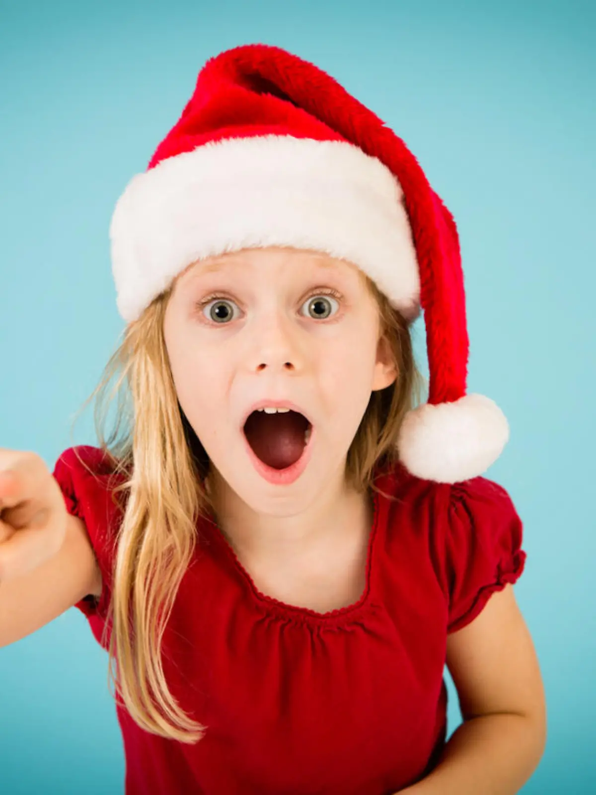 Color image of a surprised, blond-haired little girl wearing a Santa hat and pointing her finger. 