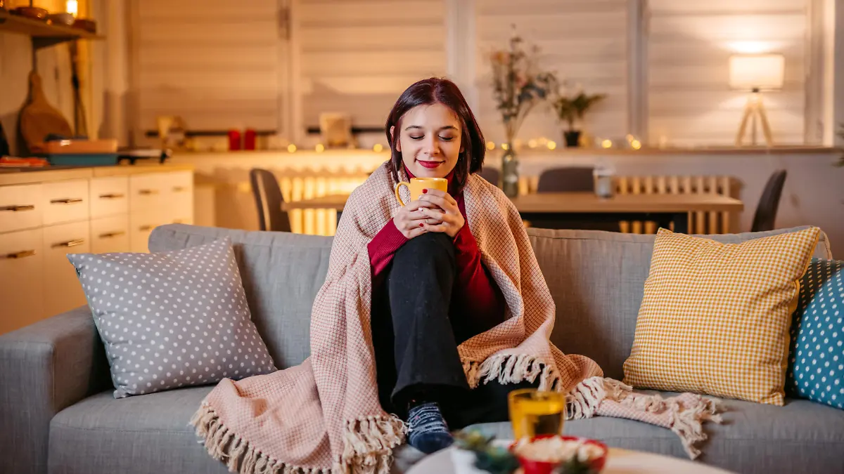 Beautiful young woman drinking hot tea wrapped in blanket on sofa at home.