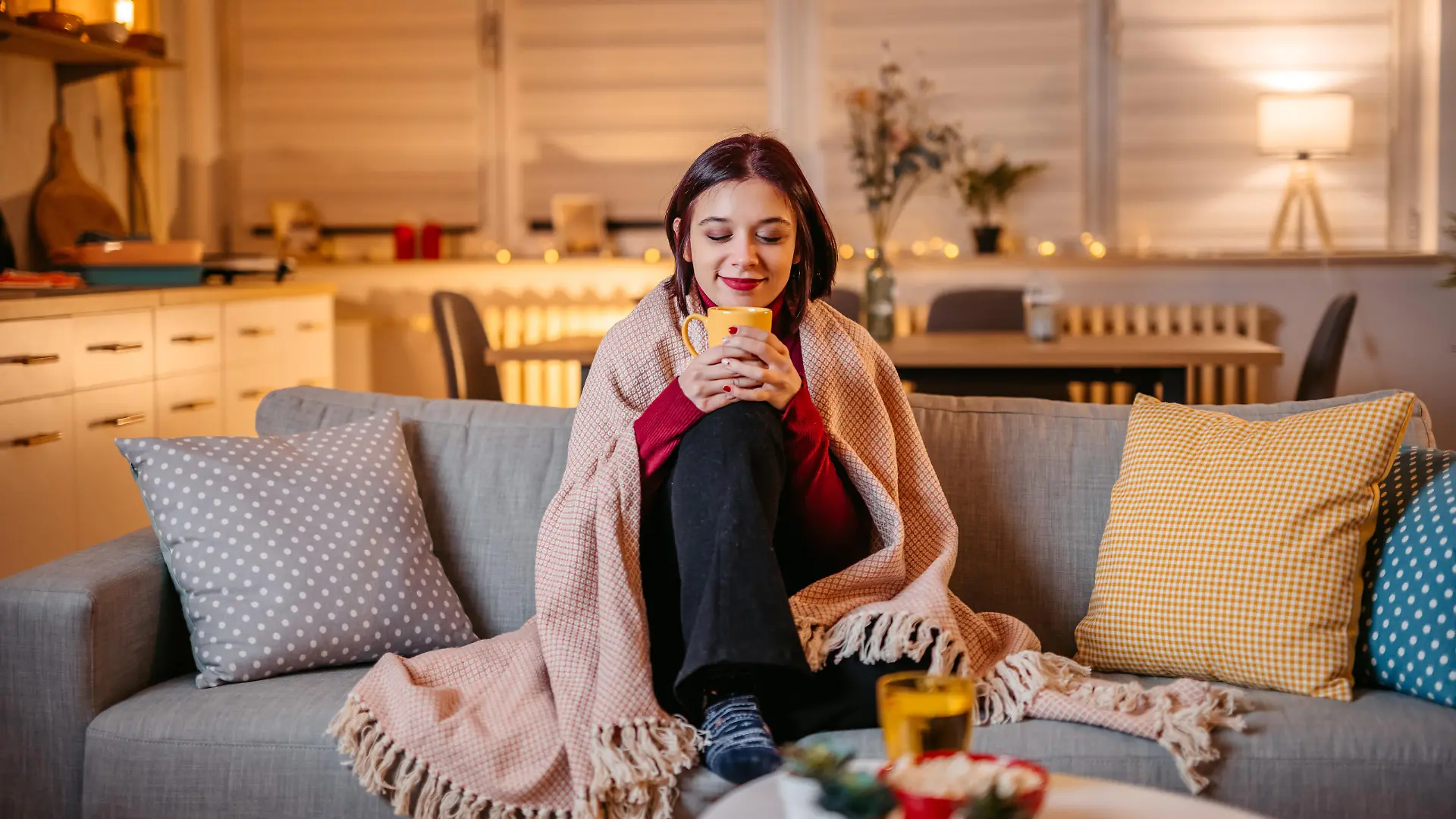 Beautiful young woman drinking hot tea wrapped in blanket on sofa at home.
