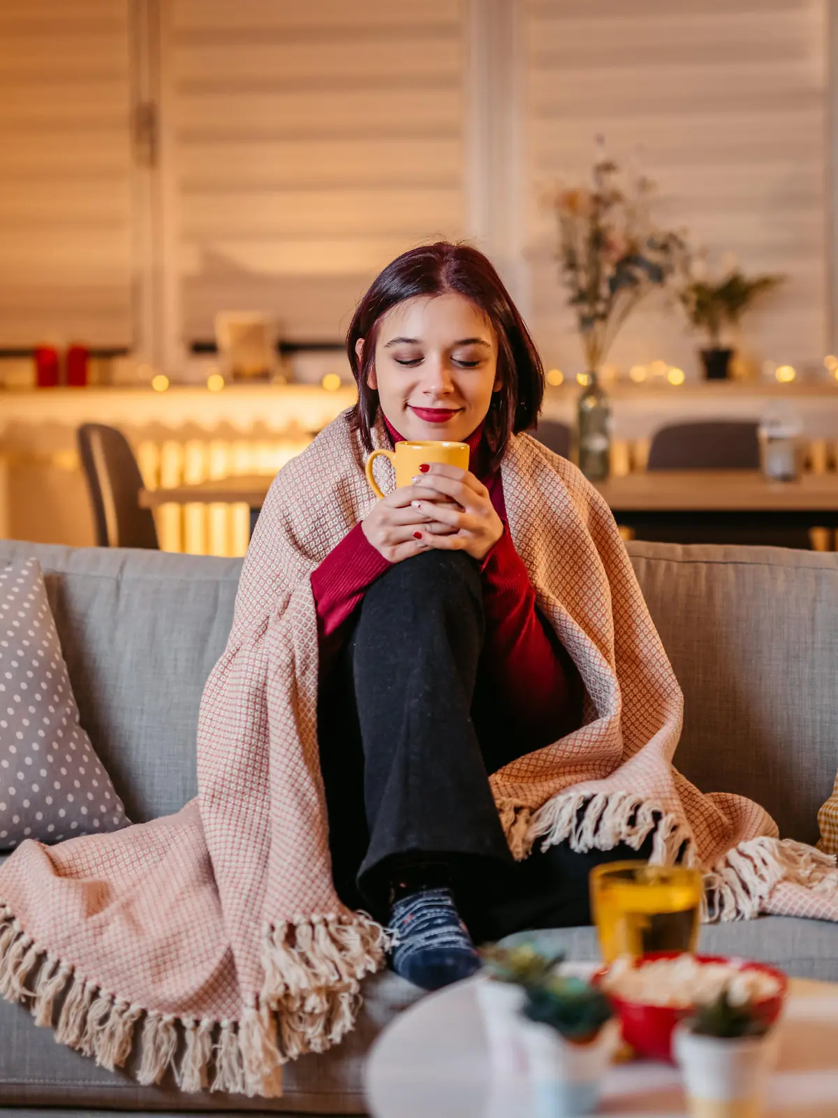 Beautiful young woman drinking hot tea wrapped in blanket on sofa at home.