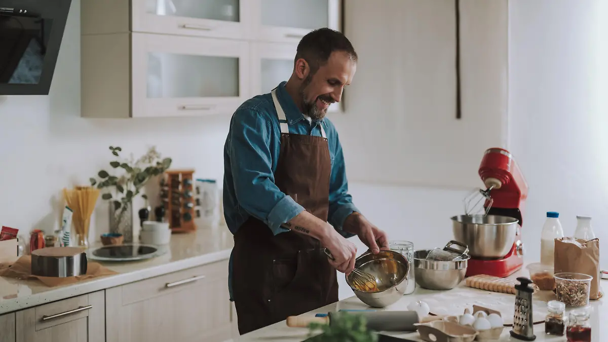 Cheerful emotional bearded man standing in the kitchen and smiling while holding a whisk and beating up eggs