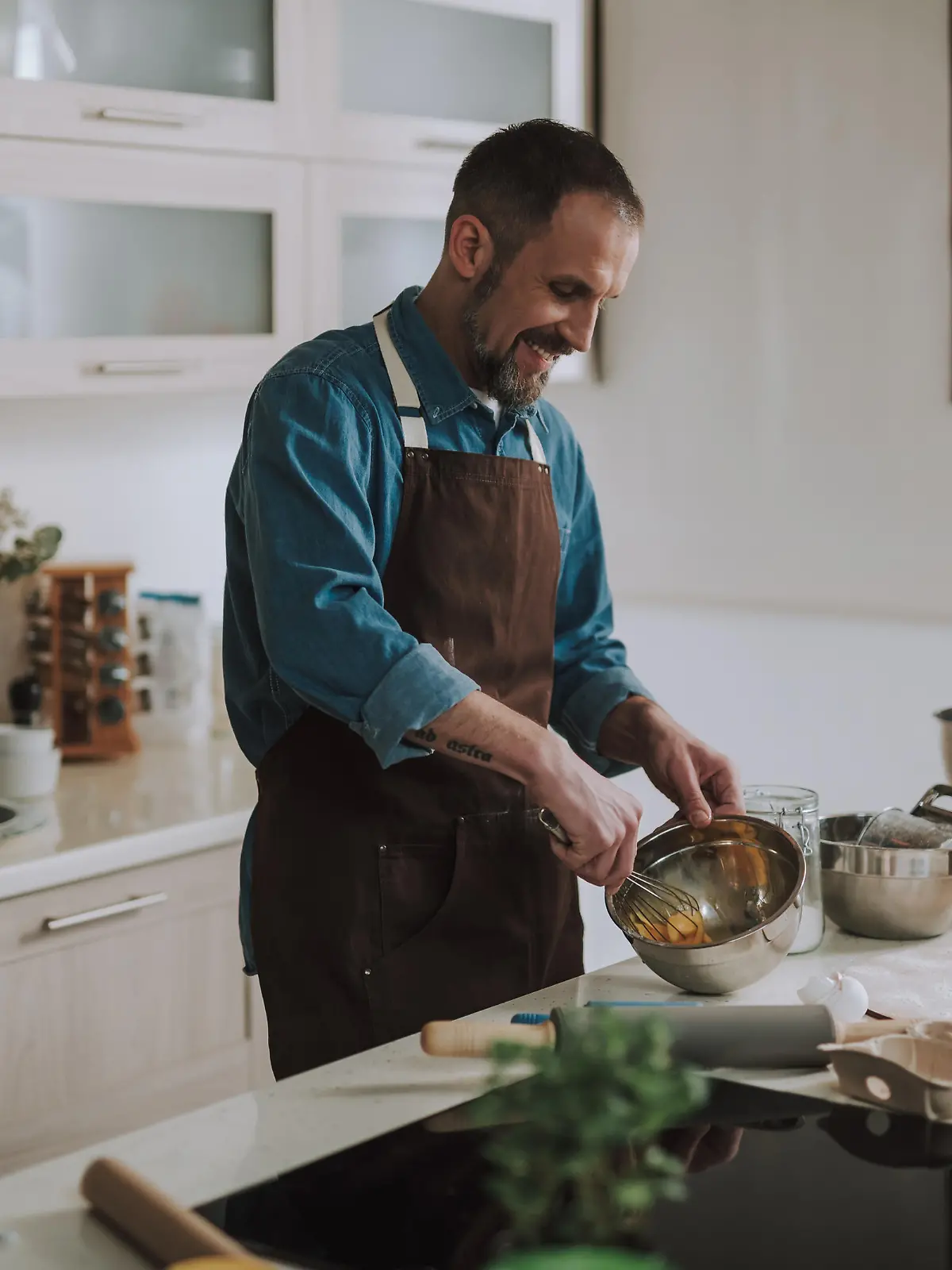 Cheerful emotional bearded man standing in the kitchen and smiling while holding a whisk and beating up eggs