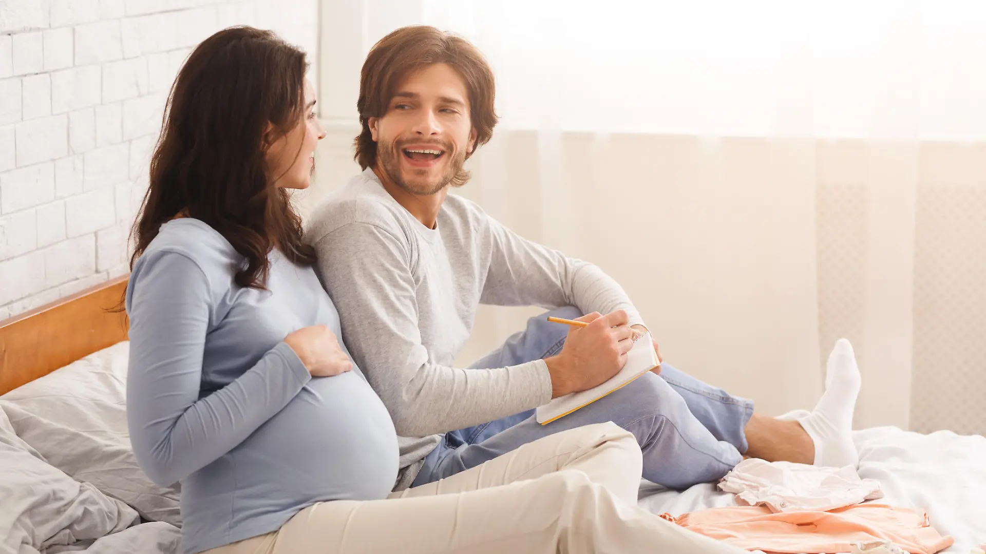 Young pregnant couple choosing name for their future baby, discussing and writing options to notebook, sitting on bed at home together, free space