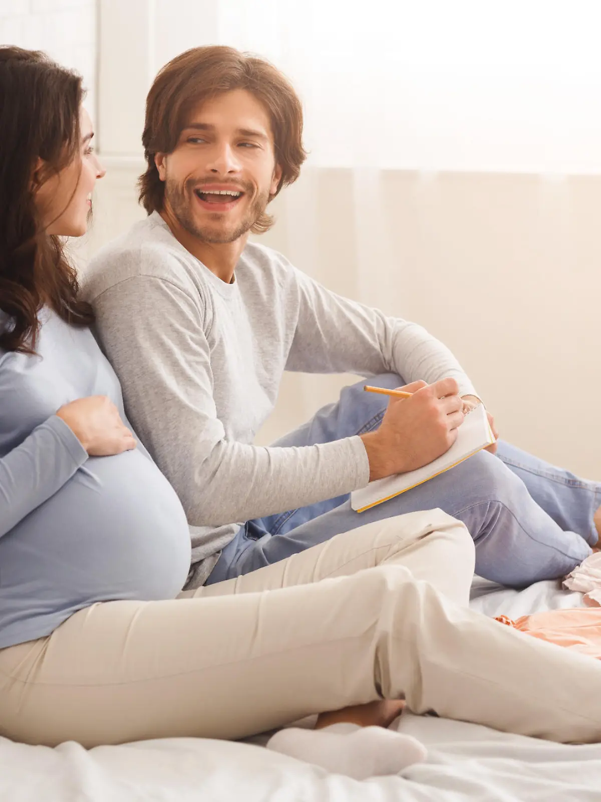 Young pregnant couple choosing name for their future baby, discussing and writing options to notebook, sitting on bed at home together, free space