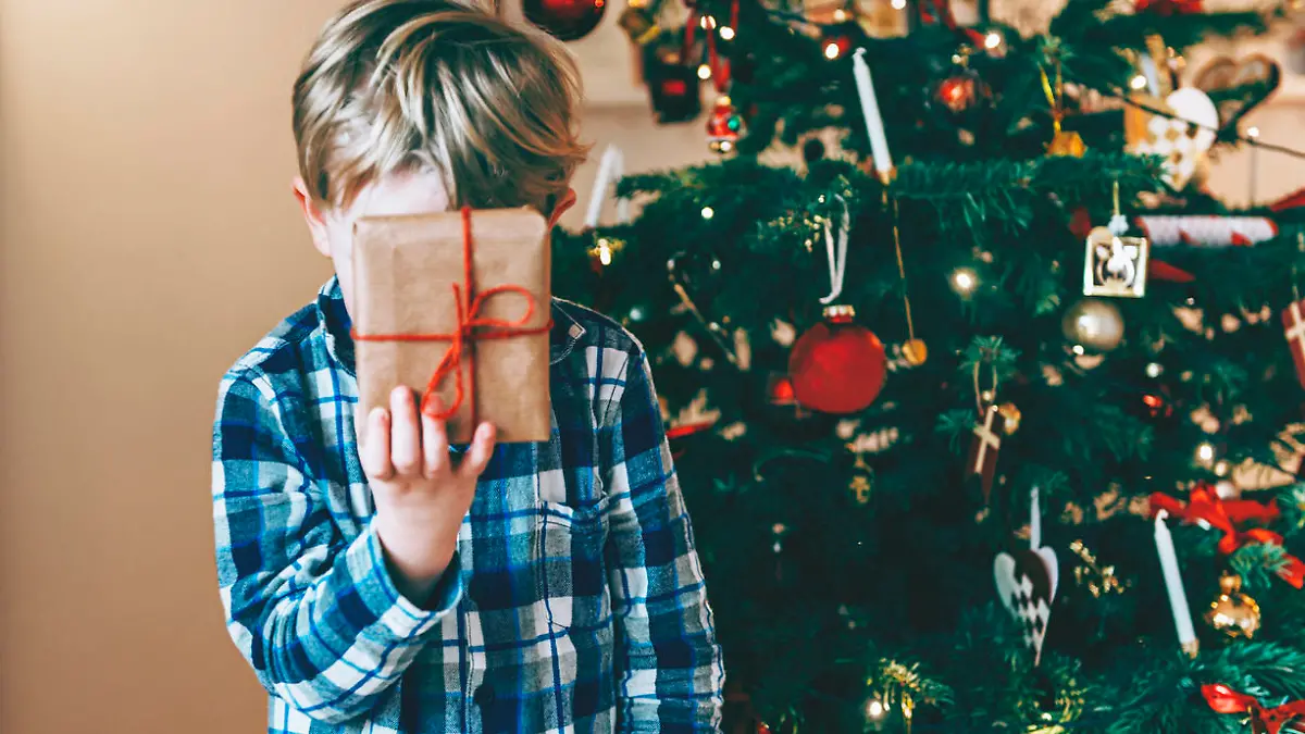 Boy holds up a present in front of his head. He has blond hair and wears a pajamas. It is christmas morning, and the child stands in front of a decorated christmas tree.