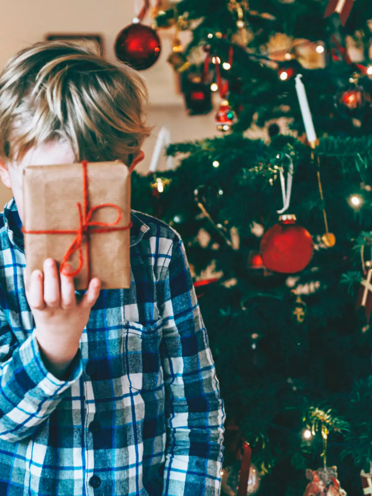 Boy holds up a present in front of his head. He has blond hair and wears a pajamas. It is christmas morning, and the child stands in front of a decorated christmas tree.