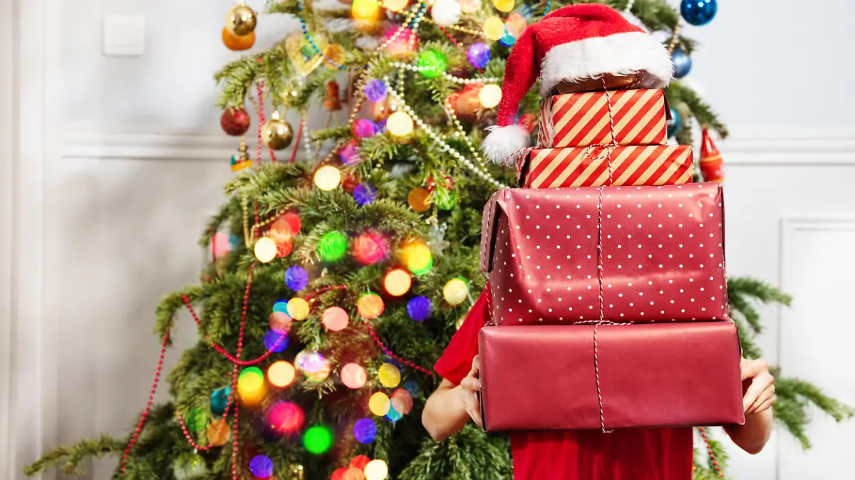Hands of a boy holding big stack of New Year presents covered with Santa hat near Christmas tree, no face visible