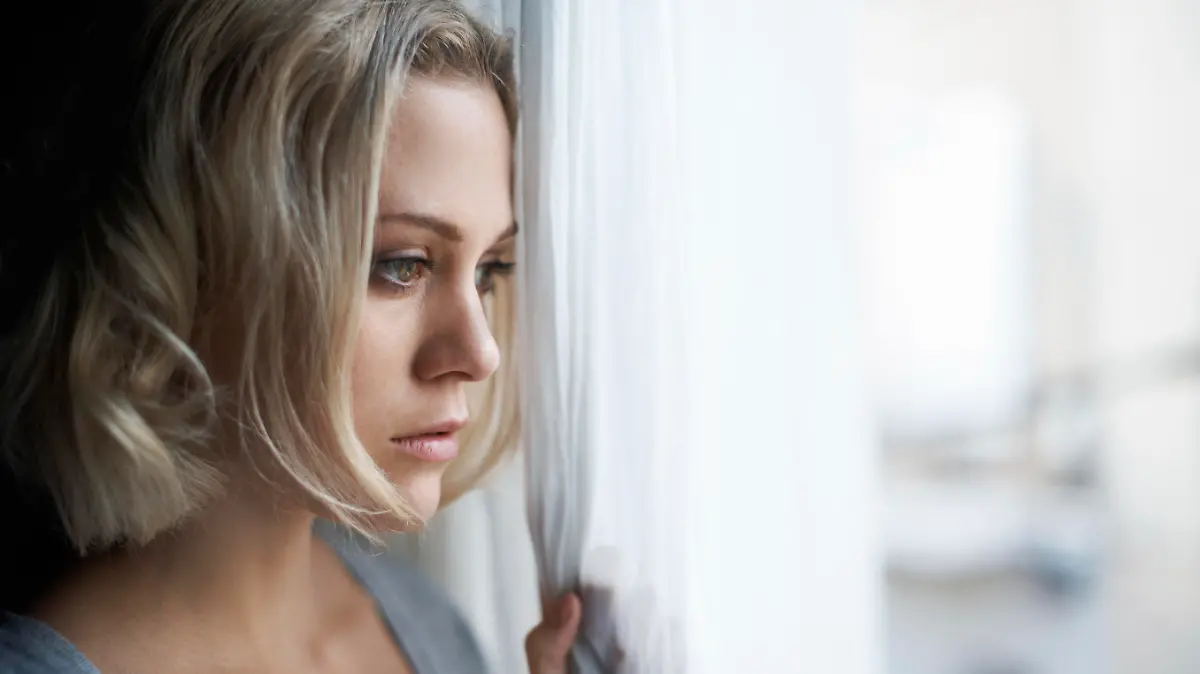 A beautiful young blonde woman looking out of her window sadly