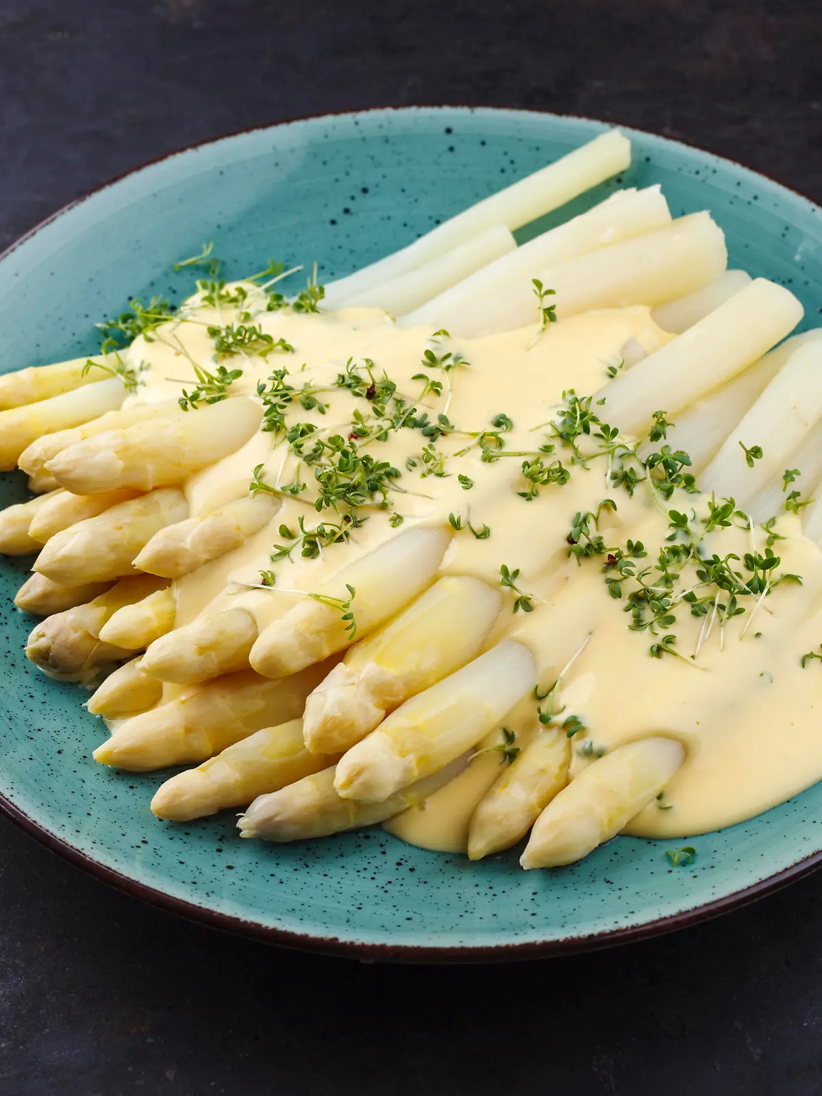 Modern style traditional steamed white asparagus with sauce hollandaise served herbs as close-up on a Nordic design plate on a rustic black board