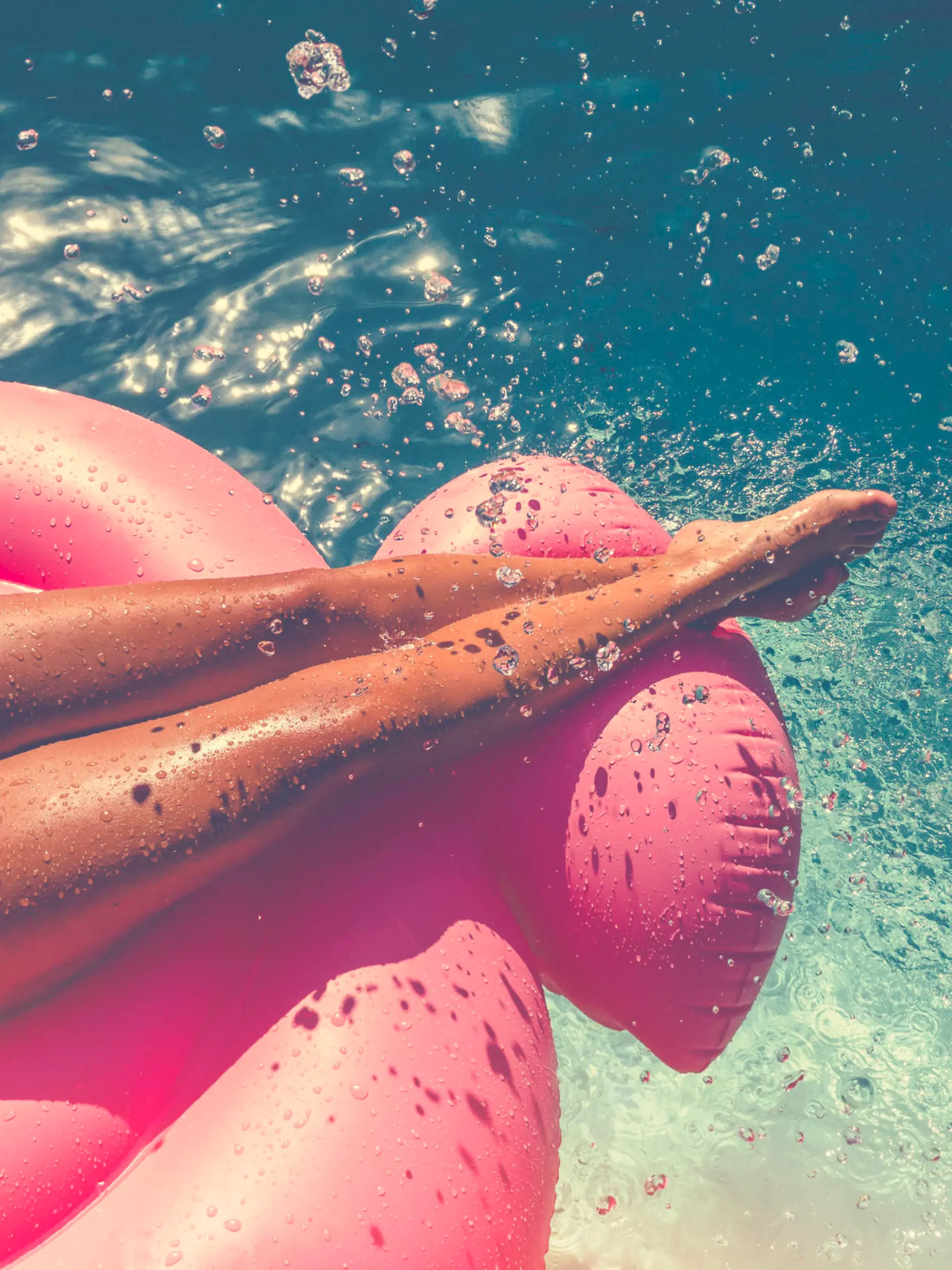 Woman floating on a pink inflatable in swimming pool. Tight crop showing only her legs and feet. She is tanned in turquoise water with water splashing onto her.