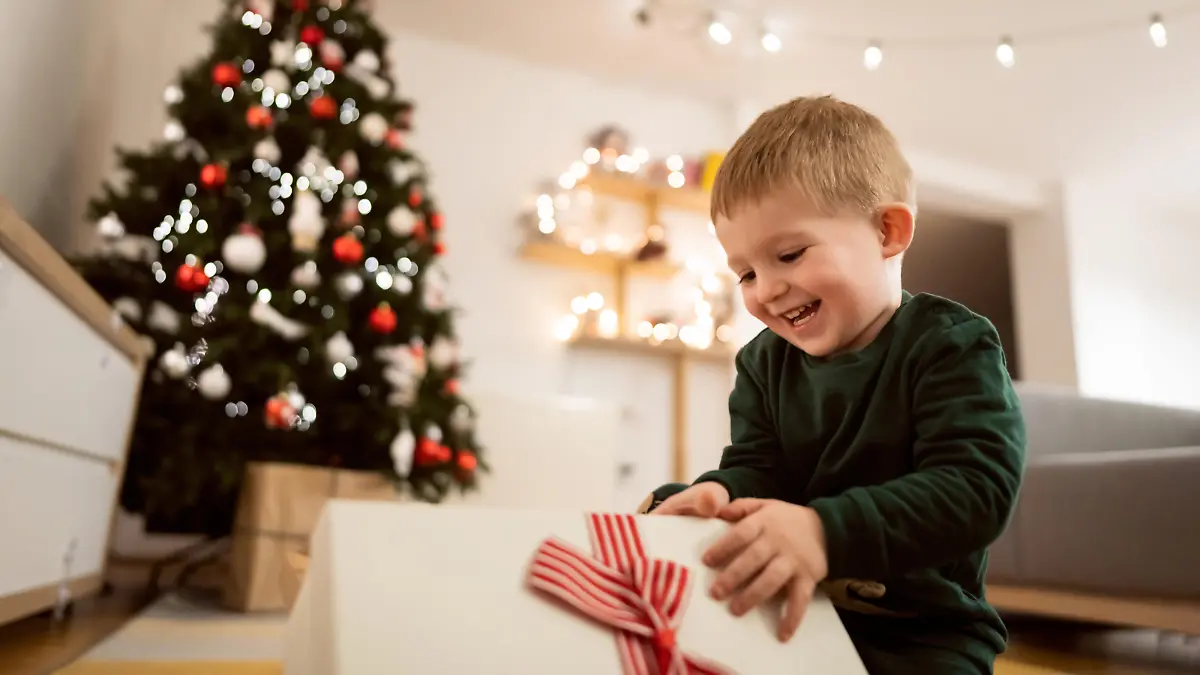 Caucasian toddler boy sitting near Christmas tree, and unwrapping his Christmas present