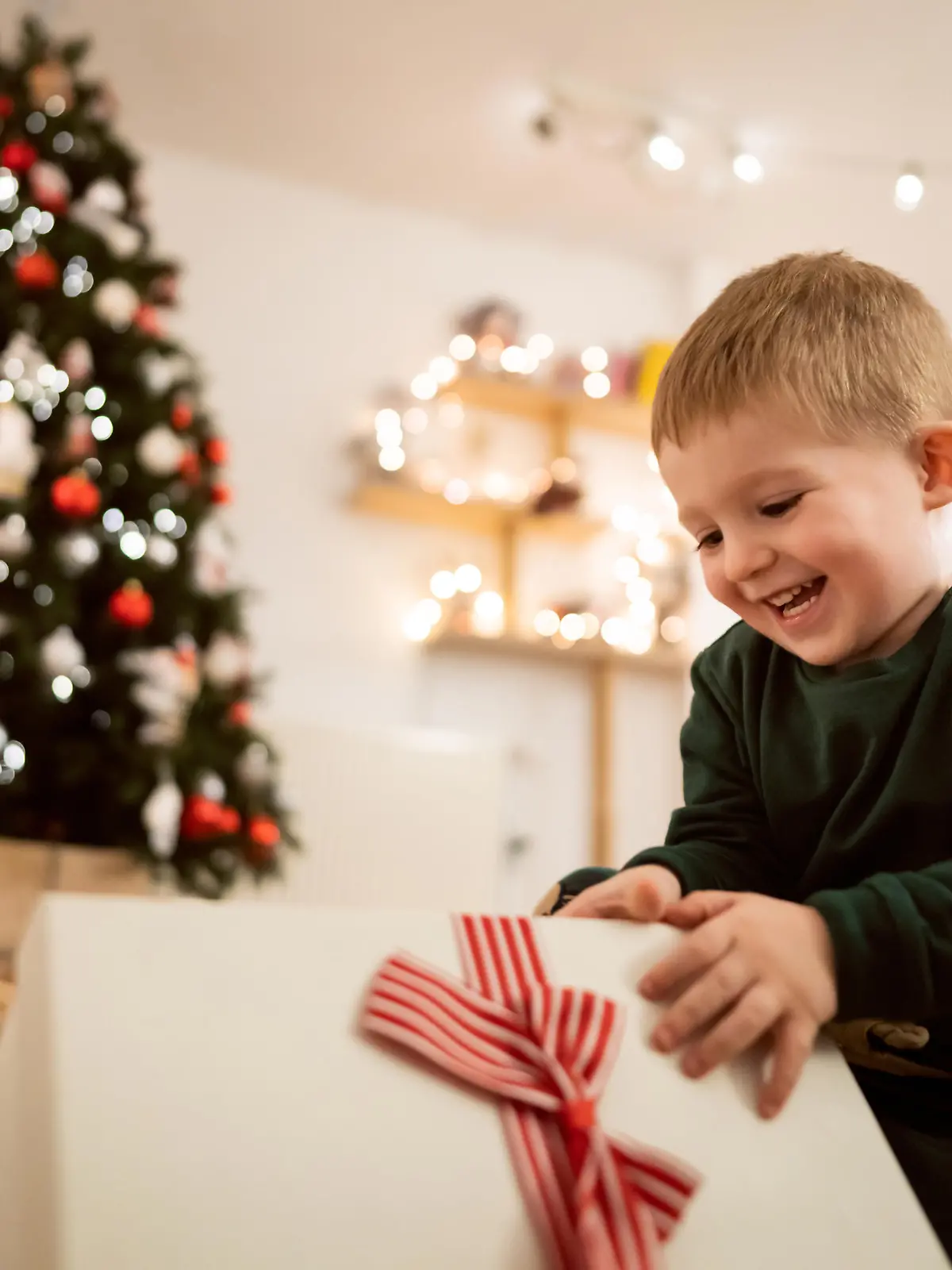 Caucasian toddler boy sitting near Christmas tree, and unwrapping his Christmas present