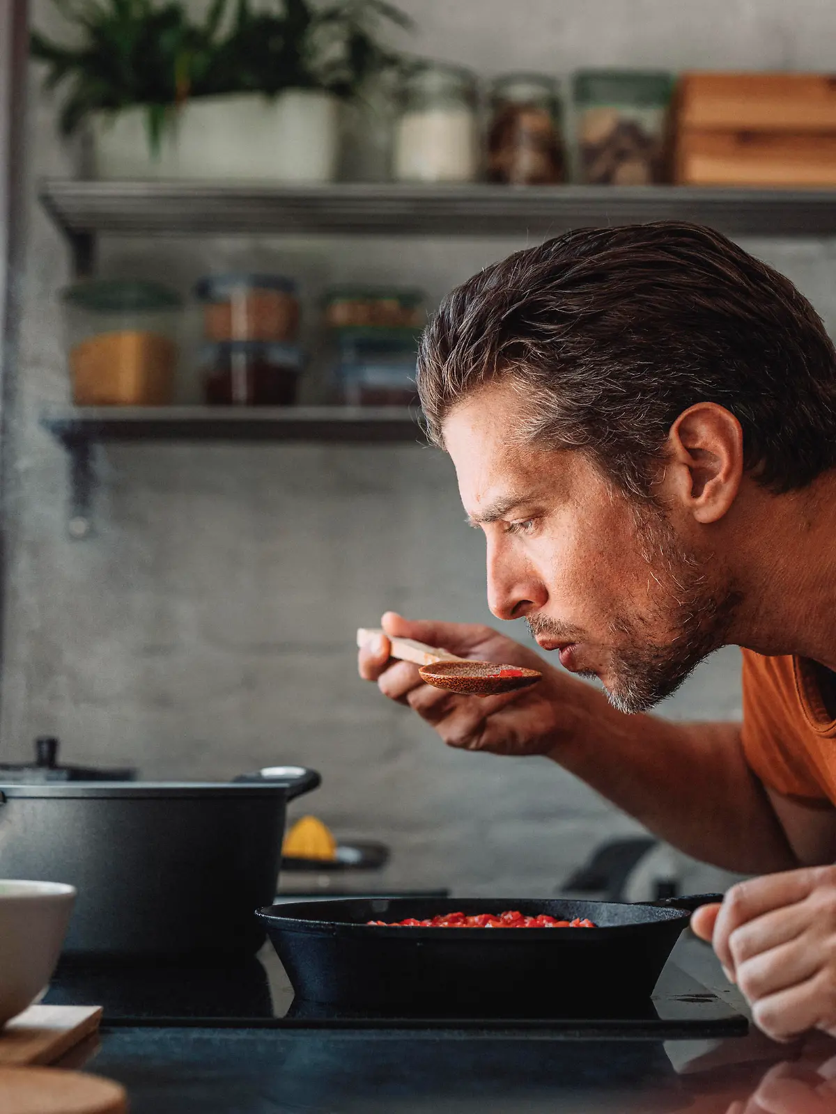 Close up shot of a handsome young happy Caucasian man tasting sauce with a mixing spoon with his eyes closed over a frying pan in a kitchen.