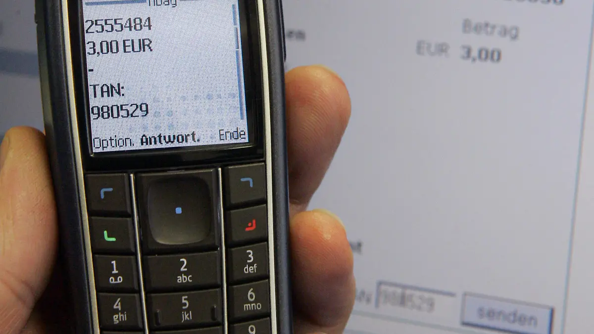 Das Symbolbild zeigt ein handelsübliches Handy mit einer Kurznachricht (SMS) mit einer gerade erzeugten TAN vor einem Monitor, auf dem eine Online-Banking-Seite der Nationalbank zu sehen ist (Foto vom 14.11.2006). Der Kunde beantragt während seines Internetbesuchs eine TAN online, bekommt diese sofort auf sein Handy geschickt, um sie dann in das entsprechende Feld am Rechner einzutragen. In den vergangenen Jahren häuften sich die Fälle des Datenklaus im Internet, dem so genannten "Phishing". Foto: Bernd Thissen dpa/lnw (zu dpa-KORR "Neue Überweisungsverfahren sollen Online-Banking sicherer machen" vom 03.12.2006) +++(c) dpa - Report+++