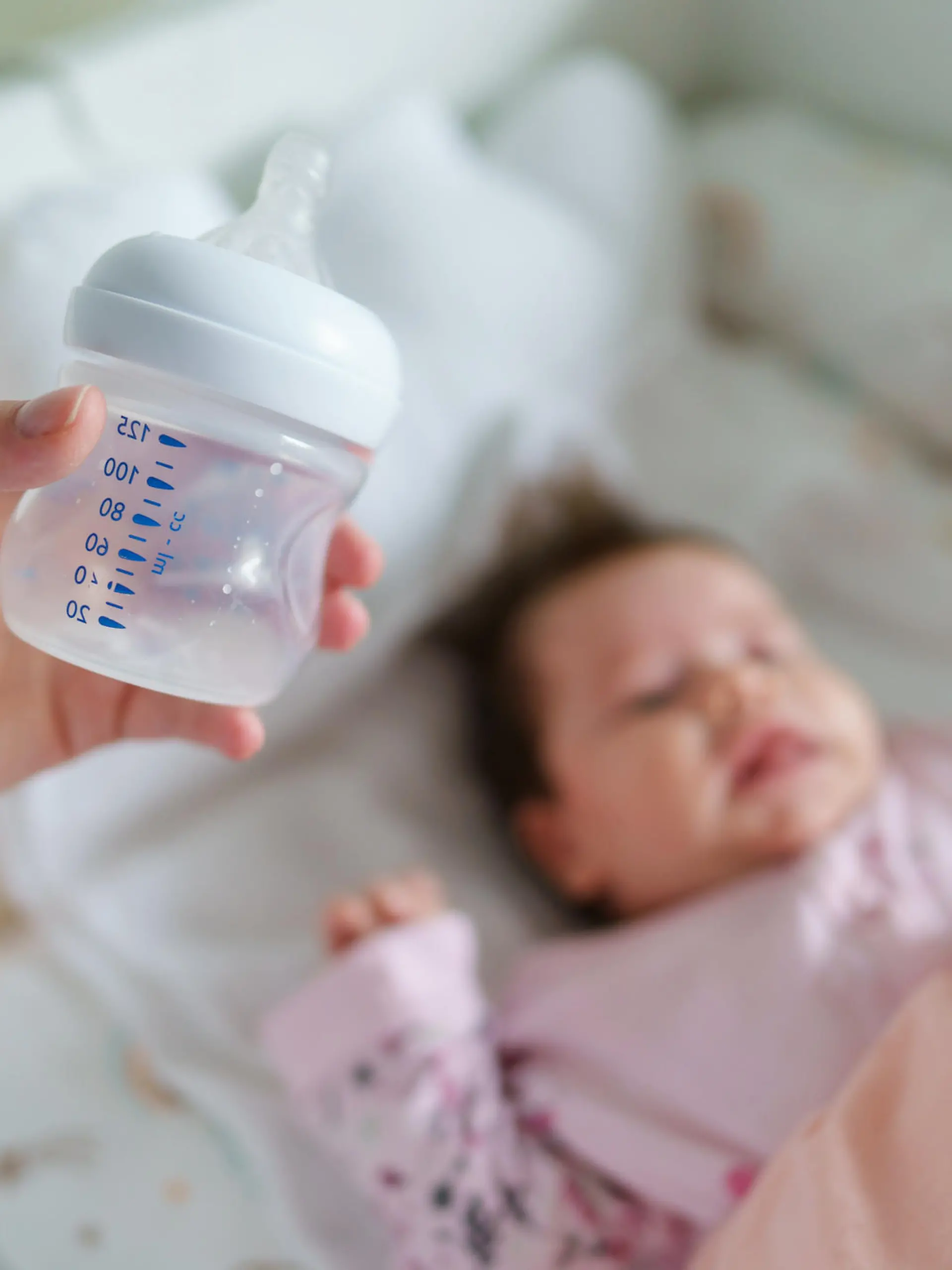 Close up selective focus on empty baby bottle in hands of unknown woman mother holding it above the bed at home with sleeping infant in background - newborn feeding nursing concept concept copy space