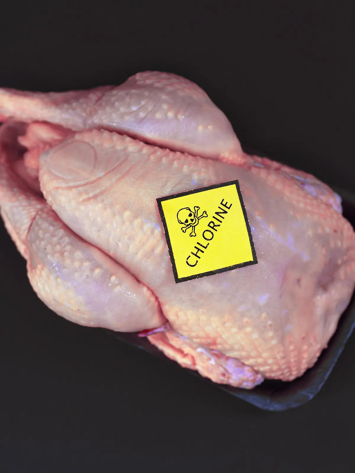 Concept for chlorinated chicken with raw whole chicken with yellow warning label with skull and word 'Chlorine' on it on black background