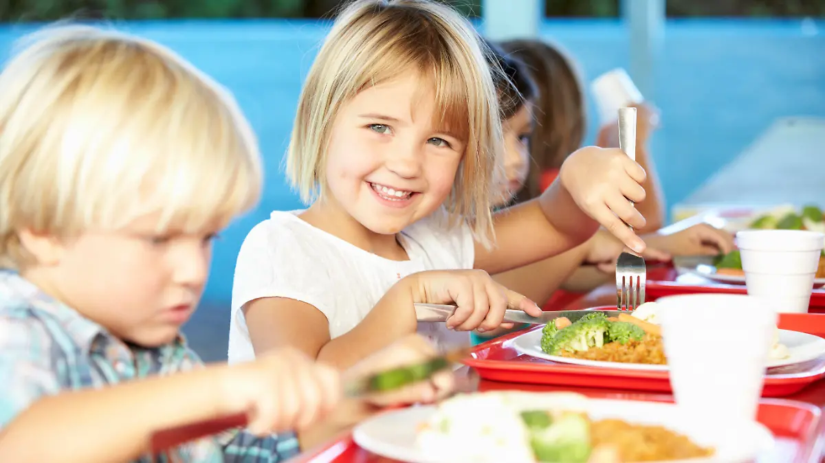 Elementary Pupils Enjoying Healthy Lunch In Cafeteria Sitting Down Smiling At Camera