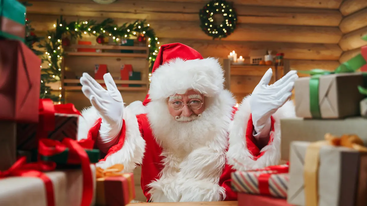 Santa Claus is  inviting us, by the gesture of his hands, we see  him in a centre of the photo  in the Christmas interior with a lot of presents in the foreground