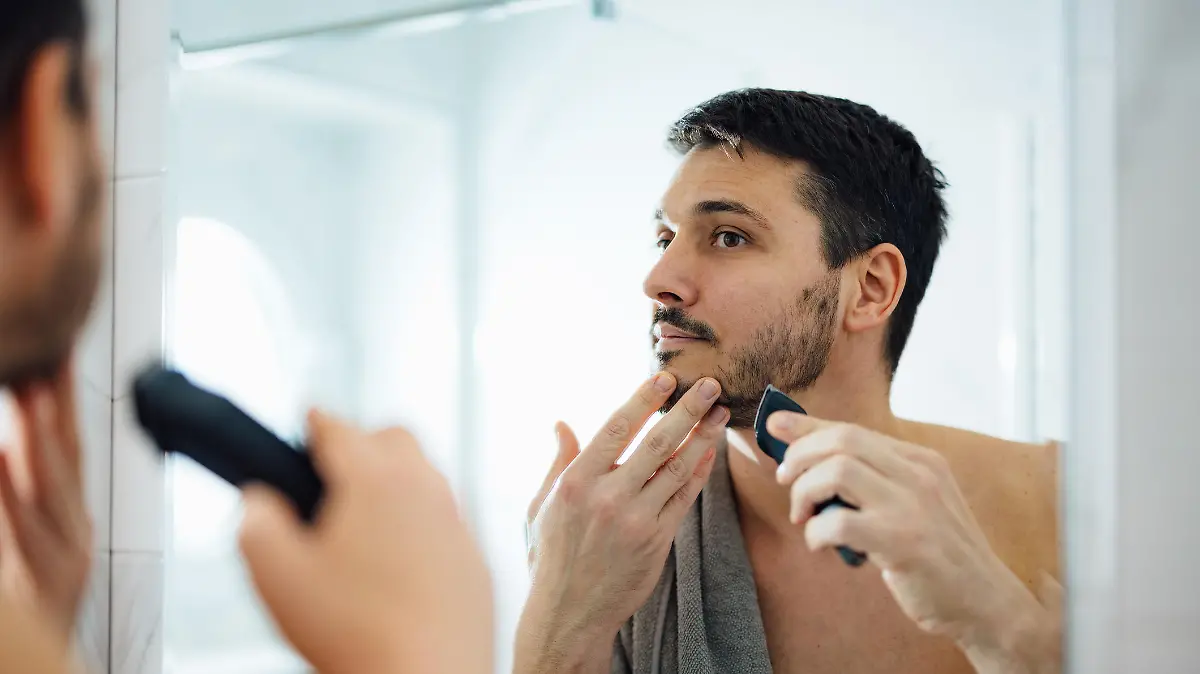 Close up photo of serious man trimming his beard with a trimmer in front of a bathroom mirror in the morning.