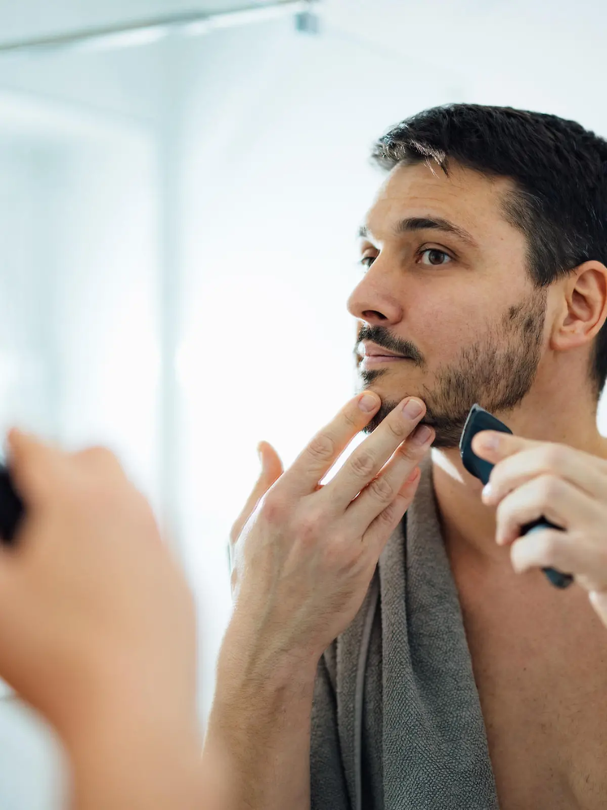 Close up photo of serious man trimming his beard with a trimmer in front of a bathroom mirror in the morning.