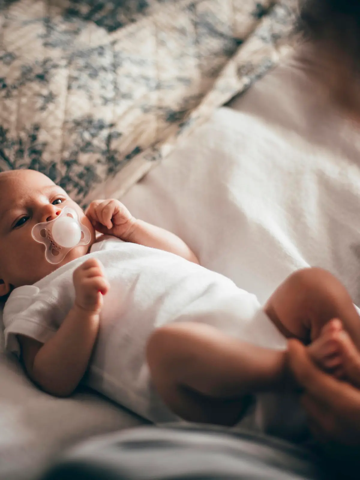 A cute little baby lies in her bed as her mother gets ready to change her nappy. She has a dummy in her mouth as she lies on a bed looking up.