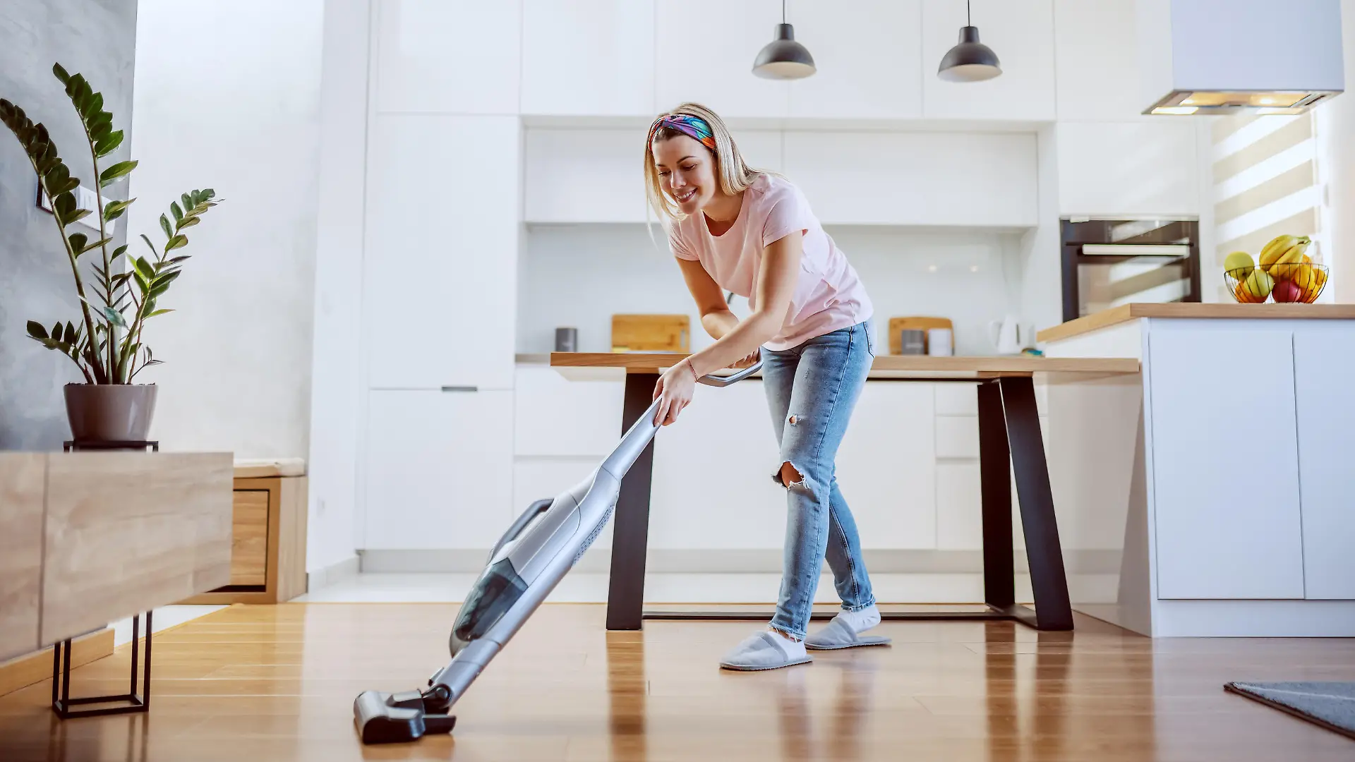 Smiling worthy Caucasian blond housewife dressed casual using steamer to clean parquet in her apartment.