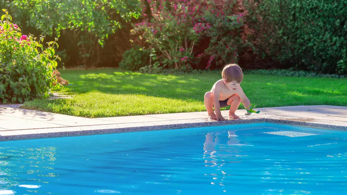 Little happy boy sitting on side of swimming pool in garden playing with his toy having fun. Summertime. Summer holidays.