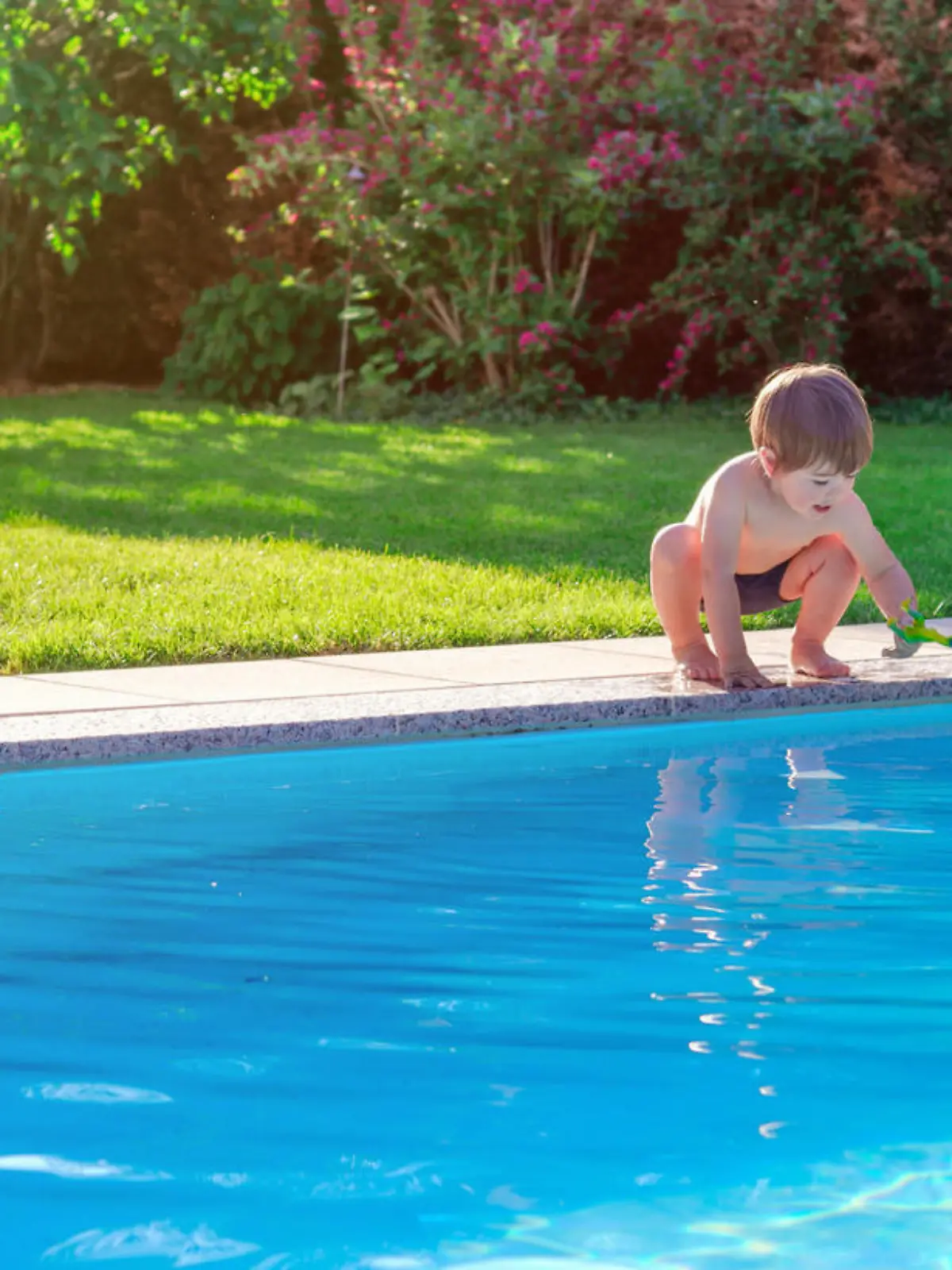 Little happy boy sitting on side of swimming pool in garden playing with his toy having fun. Summertime. Summer holidays.