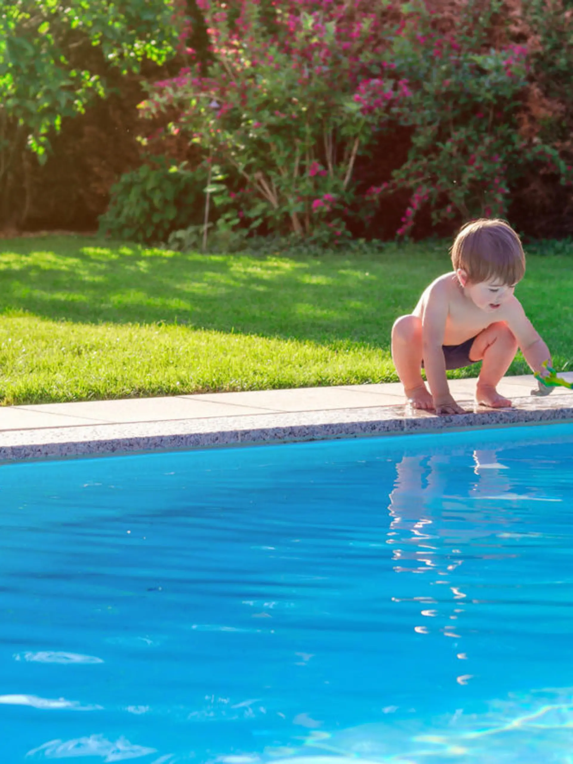 Little happy boy sitting on side of swimming pool in garden playing with his toy having fun. Summertime. Summer holidays.