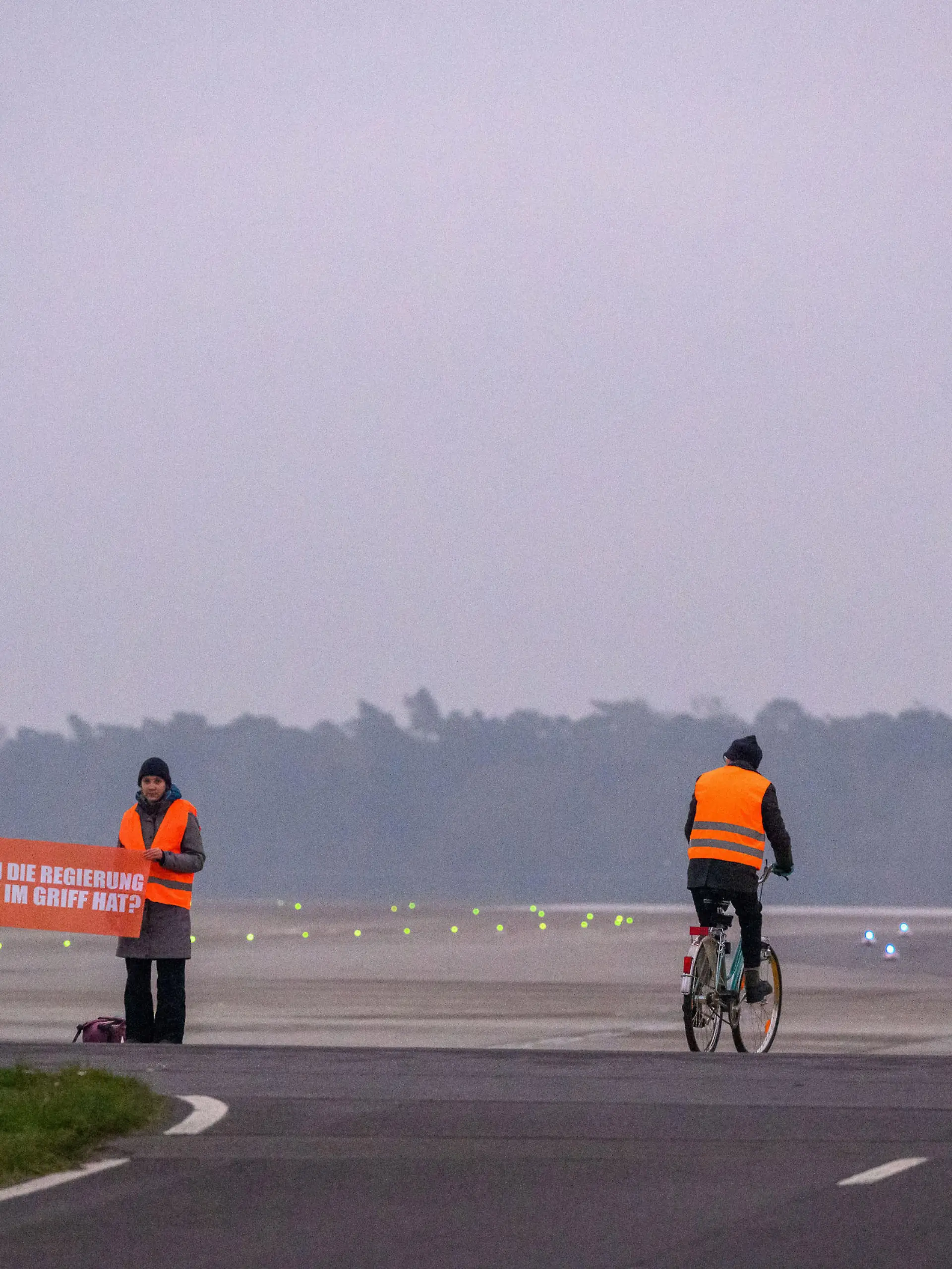 Aktivisten vom Aufstand der Letzten Generation auf der Landebahn des BER mit Fahrrad und Transparent mit der Aufschrift "Was, wenn die Regierung das nicht im Griff hat?" Schönefeld, 24.11.22