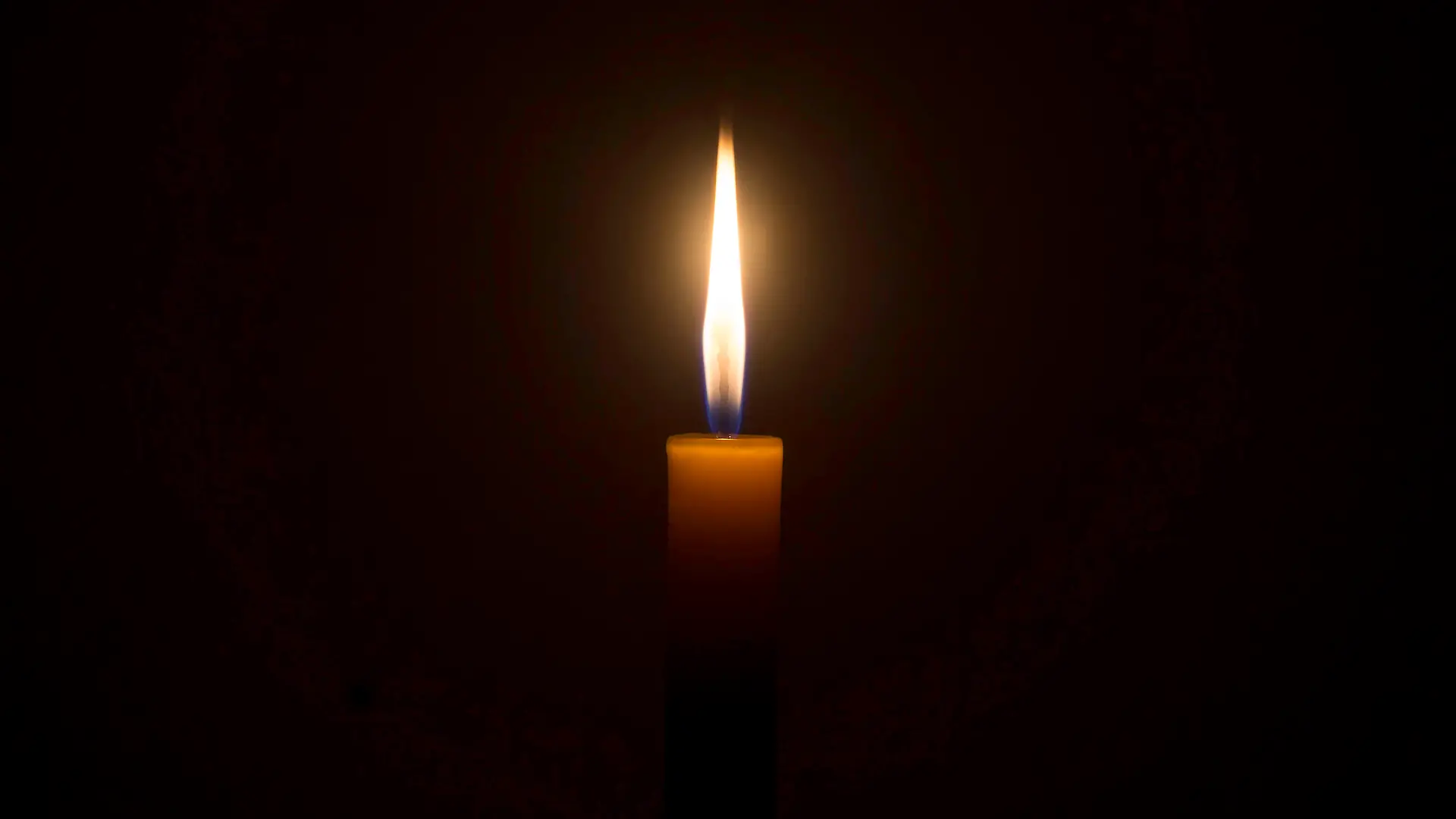 A single burning candle flame or light is glowing on an orange candle on black or dark background on table in church for Christmas, funeral or memorial service with copy space.