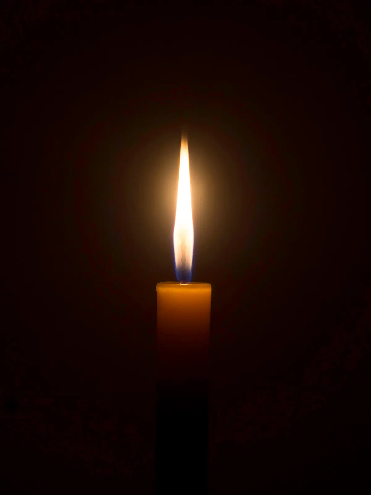 A single burning candle flame or light is glowing on an orange candle on black or dark background on table in church for Christmas, funeral or memorial service with copy space.