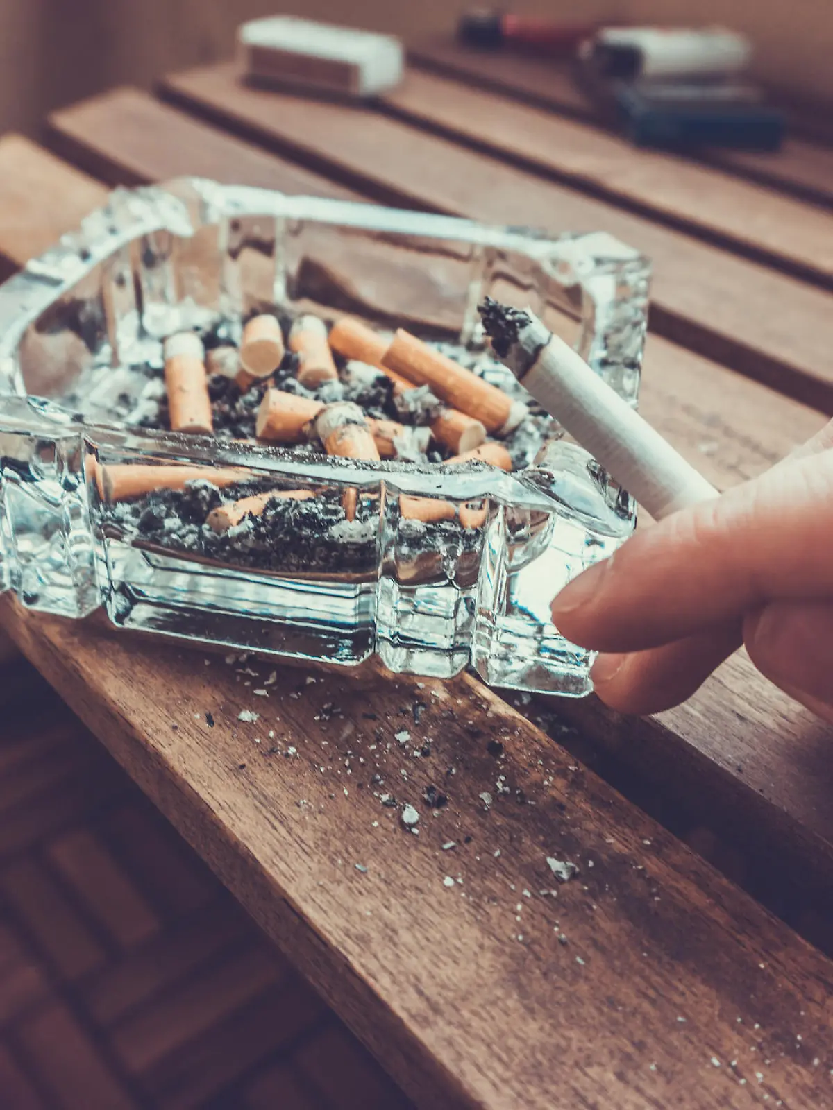 Man smoking a cigarette holding a burning filter tip in his hand alongside a glass ashtray full of ash and dead butts in a heavy smoker of addiction concept