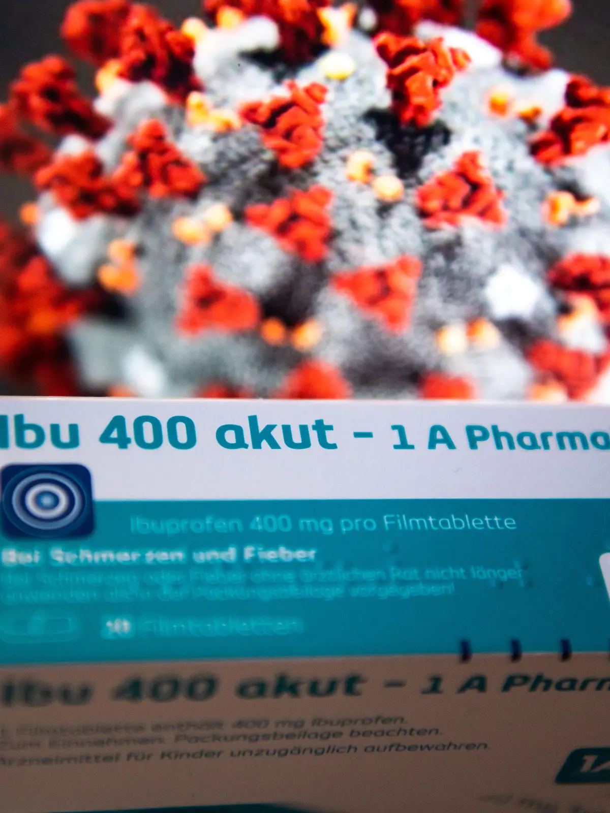 ILLUSTRATION - Eine Packung Ibuprofen liegt vor einer Abbildung des Coronavirus auf einem Tisch, die WHO hat vor der Einnahme von Ibuprofen bei einer Coronainfektion gewarnt.