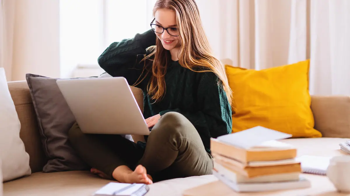 A young happy college female student sitting on sofa at home, using laptop when studying.