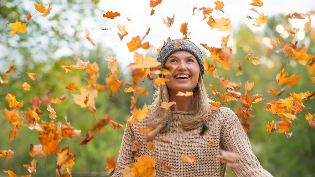 Beautiful young woman playfully smiling up at the sky as colorful autumn leaves fall around her. She is wearing a comfortable knitted sweater and a headband for warmth and is standing in a forest at sunset.