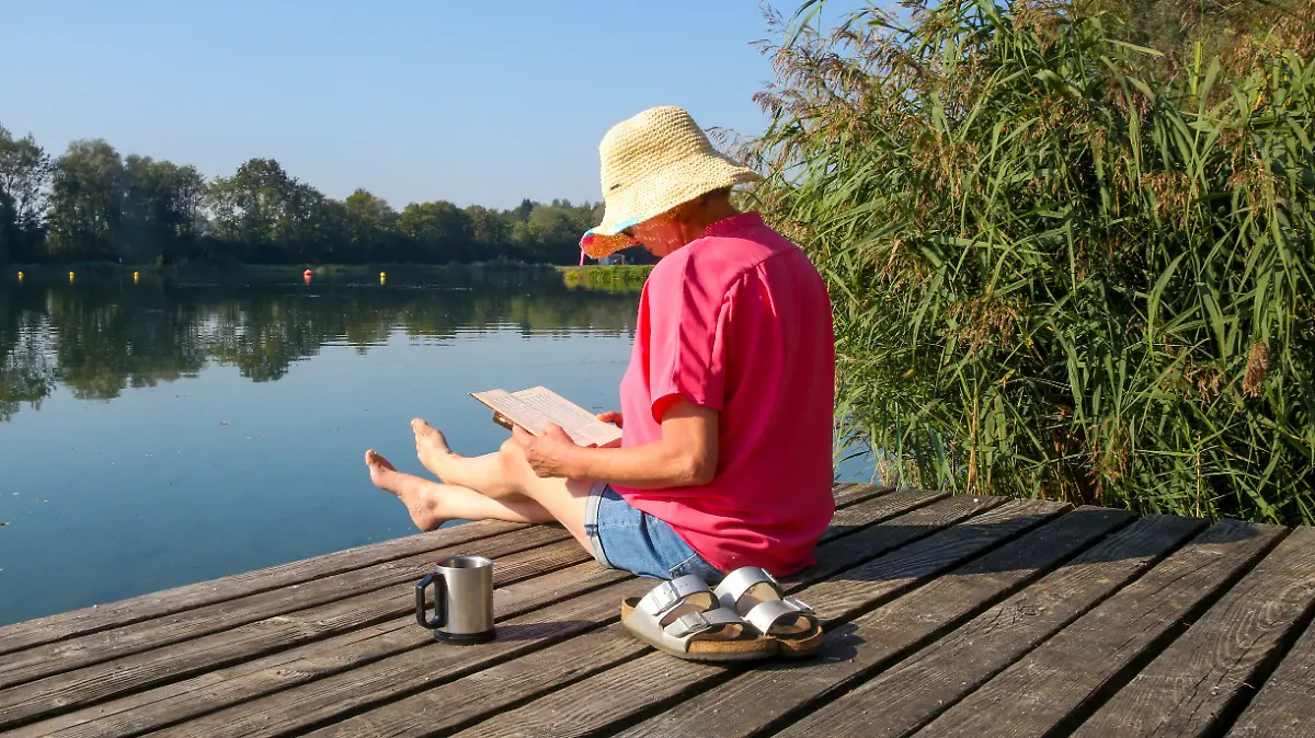 09.09.2023, Baden-Württemberg, Ertingen: Eine Frau sitzt mit einem Buch in der Hand und einer Tasse Kaffe auf einer Badeplattform in der Sonne. Foto: Thomas Warnack/dpa +++ dpa-Bildfunk +++