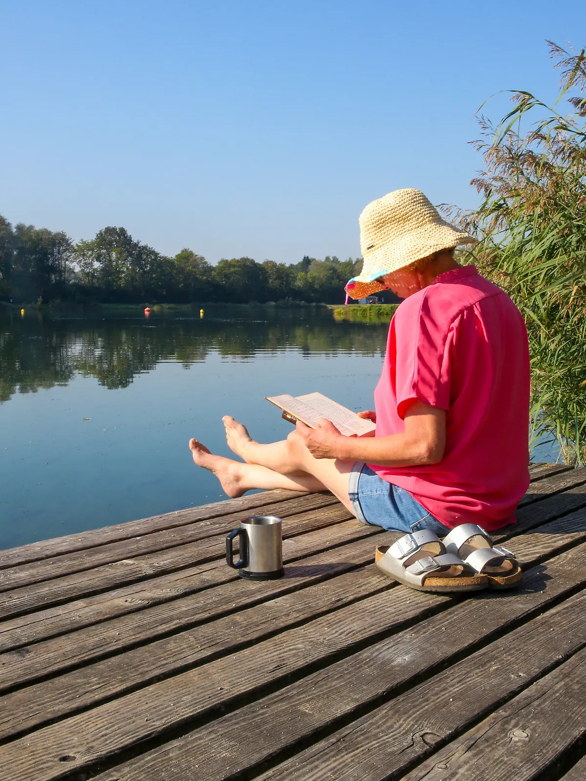 09.09.2023, Baden-Württemberg, Ertingen: Eine Frau sitzt mit einem Buch in der Hand und einer Tasse Kaffe auf einer Badeplattform in der Sonne. Foto: Thomas Warnack/dpa +++ dpa-Bildfunk +++