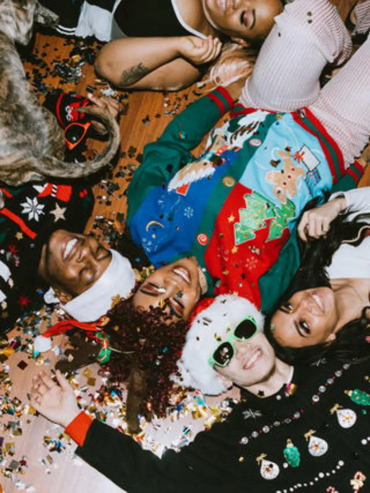 A group of young adult friends gather at a home for Christmas celebration over the holiday, dressed to fit the occasion with various Christmas accessories and ugly sweaters.  They relax on the floor, covered with confetti.